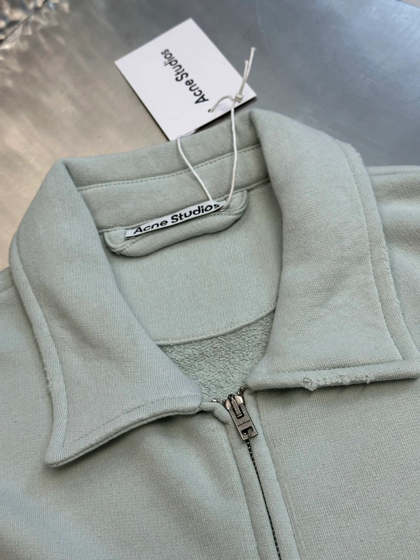 Ac vintage collared zipper sweatshirt uses 32-count top layer and 40-count bottom layer of 3-strand composite fleece structure, with a weight of g/㎡. The 32-count yarn on the top layer is fine and smooth, giving the surface a smooth touch and excellent anti-pilling properties; the 40-count 3-ply yarn on the bottom layer is treated with a raising process to form a rich and soft fluff layer, providing excellent warmth and skin-friendly wrapping. With 21 2×2 ribs, the g/㎡ weight ensures that the collar, cuffs and hem have excellent tensile fatigue resistance and shape retention capabilities. During piece dyeing, the entire fabric is immersed in the dye vat after weaving, and the dye molecules fully penetrate into the fibers, ensuring full, deep colors and excellent color fastness. Each batch of dyeing produces subtle differences, making the color tone of the single product unique. On this basis, the paint is sprayed irregularly on the surface of the garment through manual spray painting to create a splash-like retro effect. The distressed processing of holes causes the color to fade and mottle in parts. The ribbed trim is also treated to form slight whitening and fluffing, which visually echoes the spray-painted effect on the body, making the new clothes appear as if they have been worn for a long time and exposed to the sun. The front logo stamp uses a slurry printing process. The printing slurry contains a special microcapsule foaming agent. After being baked at high temperature, the slurry layer expands to form three-dimensional bulges, and the surface presents a granular texture similar to inkjet paint. This process simulates the effect of random drops of pigment after being atomized during spray gun spraying. The edges of the pattern present a natural feathering transition, rather than the rigid boundaries of glue printing. The printing surface has a fine graininess and soft elasticity to the touch, forming a closed loop of the process with the distressed effect of the spray painting on the body. size：xs s m l